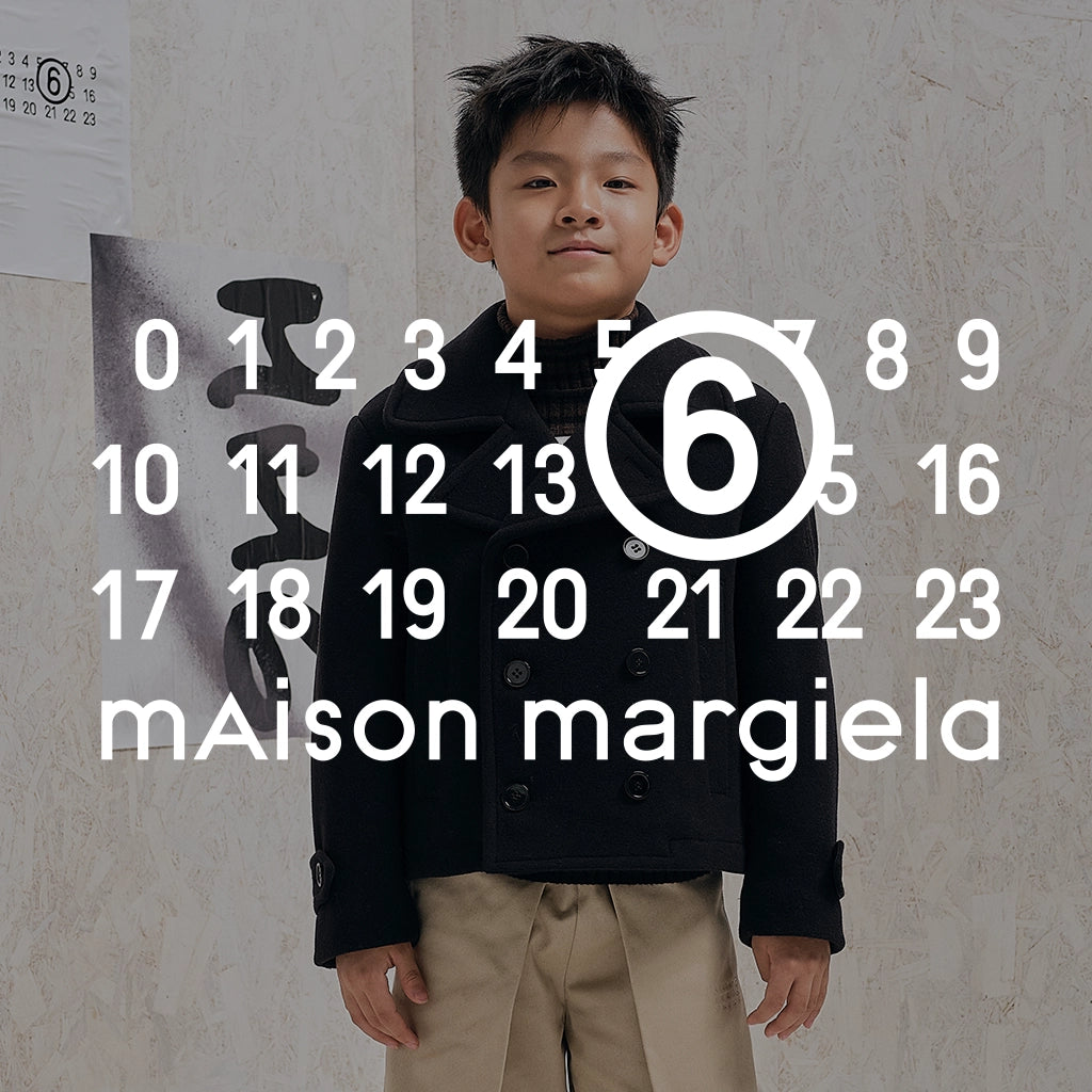 A young boy wearing a black double-breasted jacket and beige trousers stands in front of a textured wall with a large white calendar-style number overlay reading 6, with the Maison Margiela logo at the bottom.
