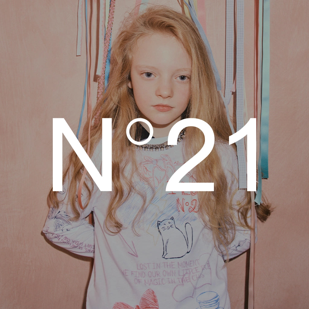 A young girl with long blonde hair looks directly at the camera, wearing a white graphic t-shirt with a cat illustration and text, set against a pinkish-beige background with hanging colorful ribbons. Large white text 'N°21' is overlaid across the center.